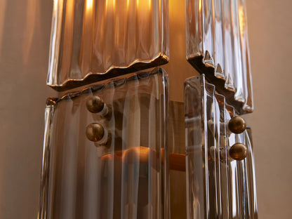 Delsie Double Sconce, vertical, burnished brass/bronze with clear glass rods & two bulbs, on a light wall next to a mirror.