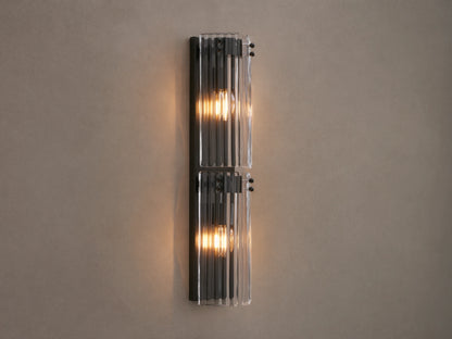 Vertical Delsie Double Sconce, dark frame with two illuminated bulbs behind ribbed clear glass, mounted on a wall near a mirror.