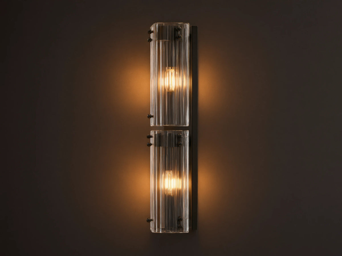 Vertical Delsie Double Sconce, dark frame with two illuminated bulbs behind ribbed clear glass, mounted on a wall near a mirror.