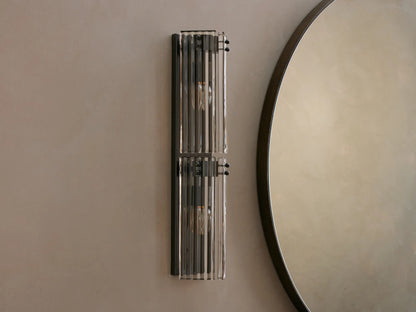 Delsie Double Sconce with two illuminated bulbs and fluted glass shades, mounted vertically on a light wall next to a round mirror.