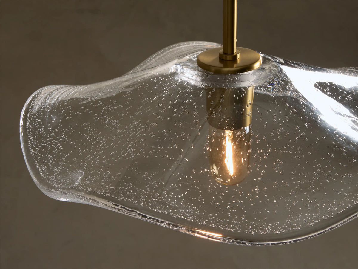 Close-up of a clear glass pendant light with an organic wavy shade, featuring internal seed glass bubbles and a brass-finished rod housing an exposed Edison bulb.
