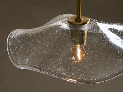 Close-up of a clear glass pendant light with an organic wavy shade, featuring internal seed glass bubbles and a brass-finished rod housing an exposed Edison bulb.