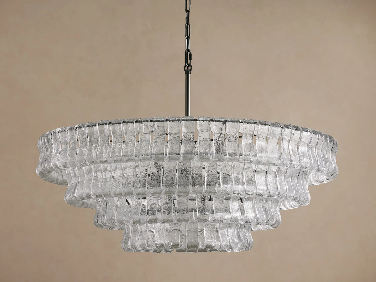 Ghiaccio Round Chandelier, multi-tiered with clear, textured curved glass pieces, hanging from a dark chain against a beige wall.