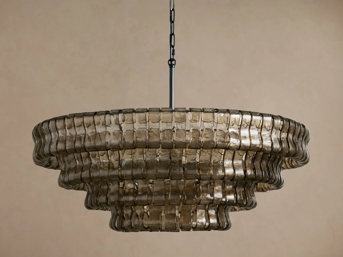 Tiered Ghiaccio Round Chandelier made of smoky, textured curved glass, suspended by a dark chain against a beige wall.