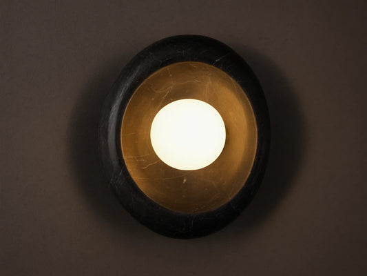 Close-up of a round black cracked marble wall sconce with a distressed gold interior and a glowing white spherical bulb.
