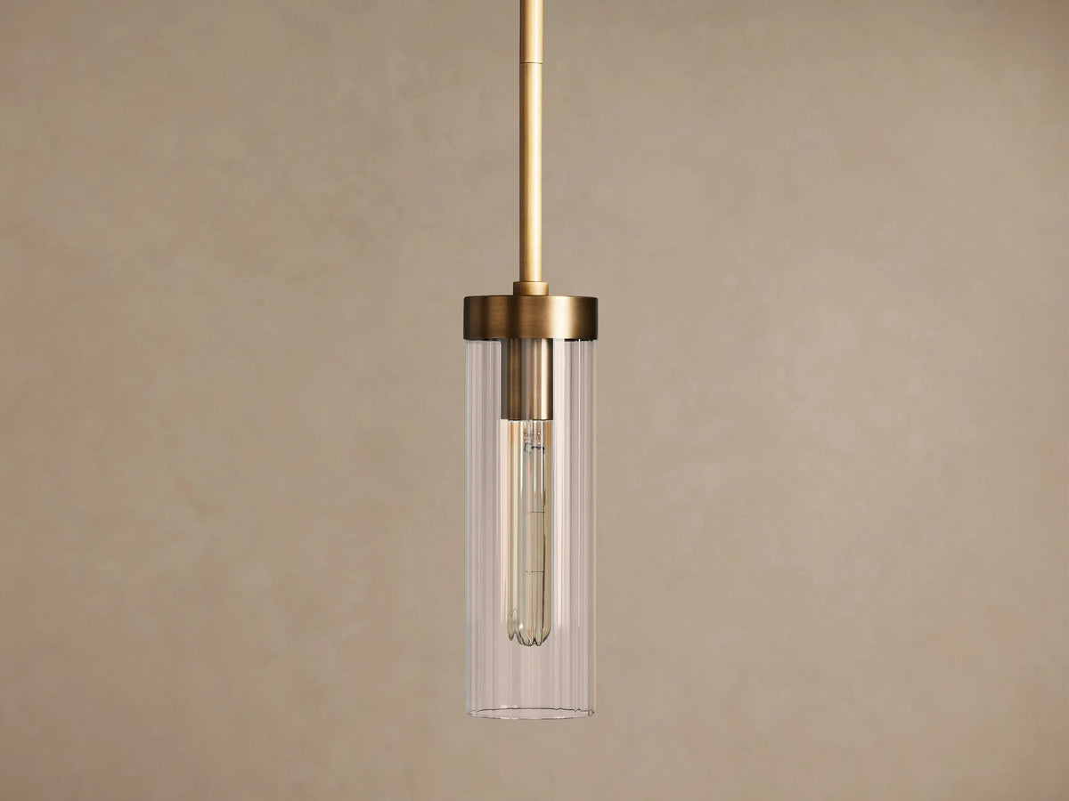 Brass Andreas Pendant with clear ribbed glass shade & illuminated filament bulb, hanging vertically against a neutral wall.