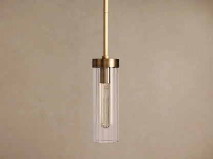 Brass Andreas Pendant with clear ribbed glass shade & illuminated filament bulb, hanging vertically against a neutral wall.