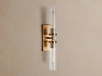 Andreas Double Sconce: rectangular brass backplate, two glowing T9 bulbs inside clear ribbed glass cylinders, mounted on a warm grey wall.