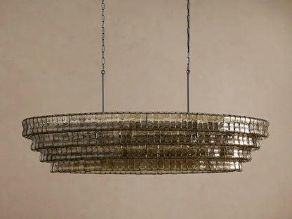 Ghiaccio Oval Chandelier, tiered with textured, curved smoke glass, glowing warmly, suspended by chains against a neutral wall.