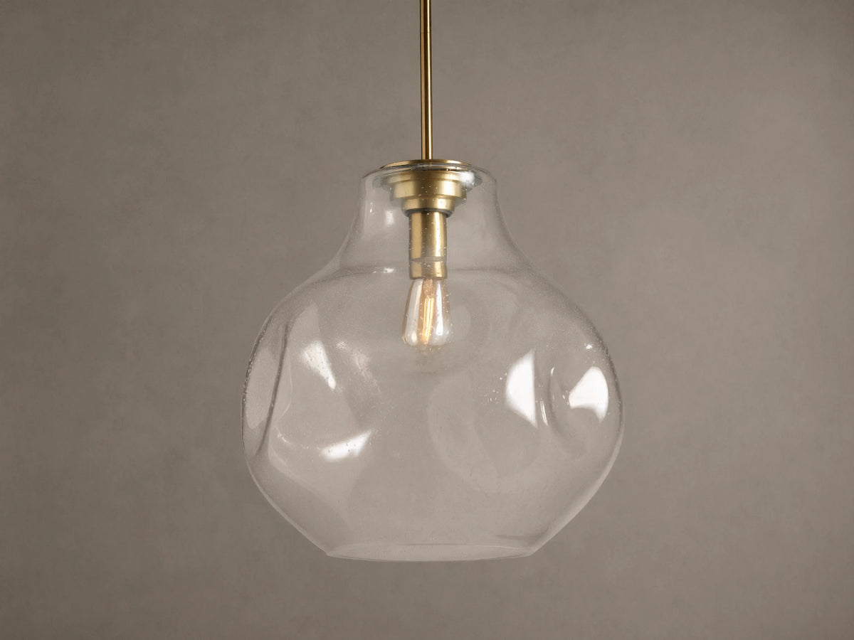 Organic 16" Pendant: Clear, seeded mouth-blown glass shade, brass stem, glowing filament bulb, against a brown wall.