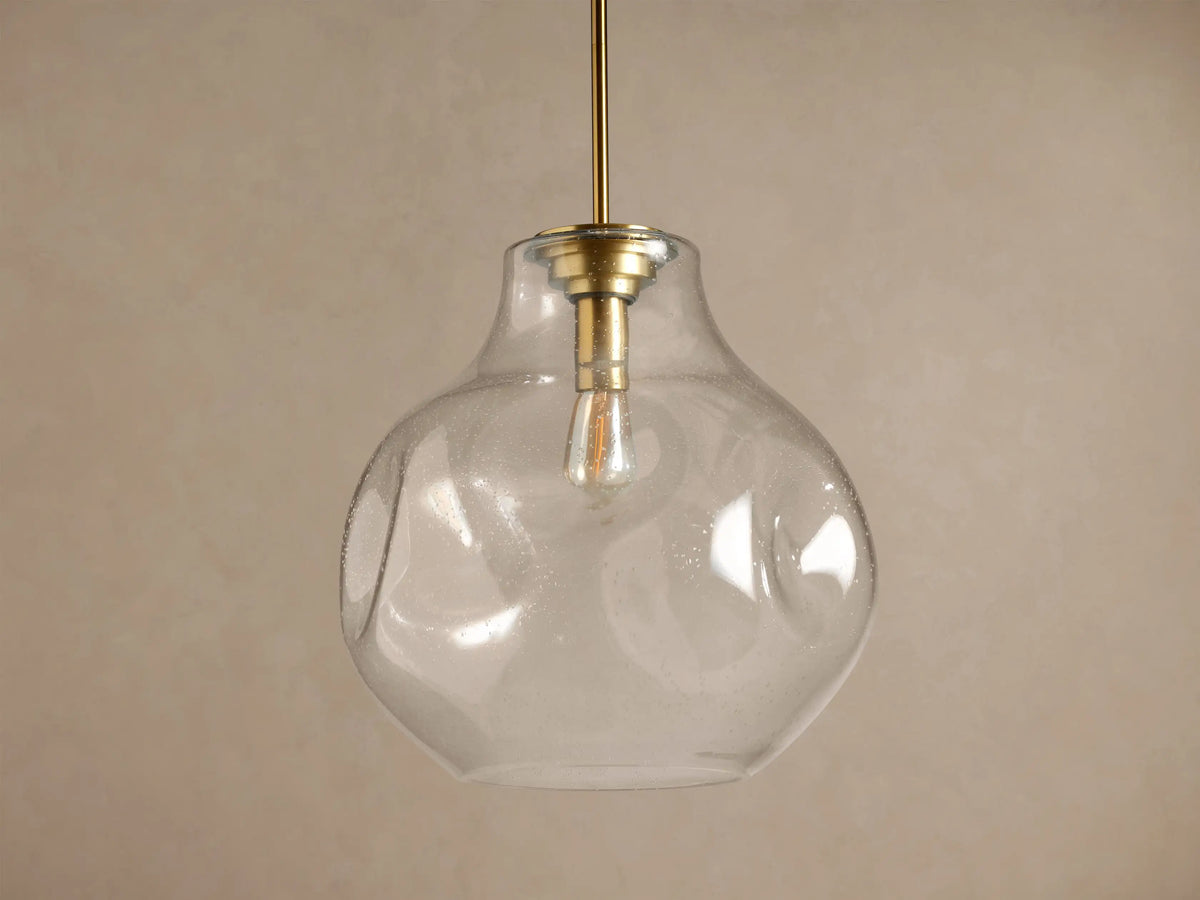 Organic 16" Pendant: Clear, seeded mouth-blown glass shade, brass stem, glowing filament bulb, against a brown wall.
