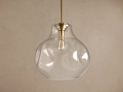 Organic 16" Pendant: Clear, seeded mouth-blown glass shade, brass stem, glowing filament bulb, against a brown wall.
