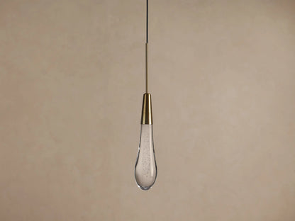 Soltaire Pendant: illuminated clear glass teardrop with internal bubbles & brass neck, hanging from a black cord against a neutral wall.