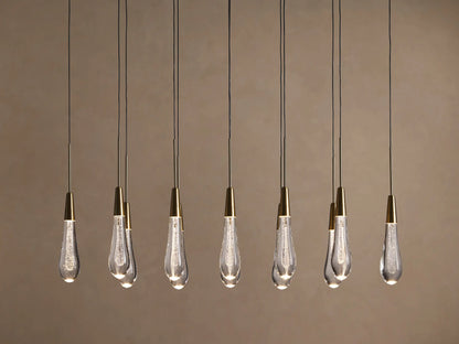 Soltaire Linear Chandelier with 8 clear, tear-drop glass pendants featuring brass tops & internal bubbles, glowing warmly.