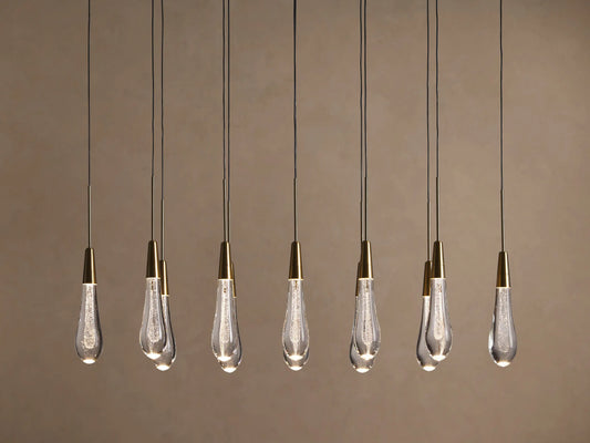 Soltaire Linear Chandelier with 8 clear, tear-drop glass pendants featuring brass tops & internal bubbles, glowing warmly.