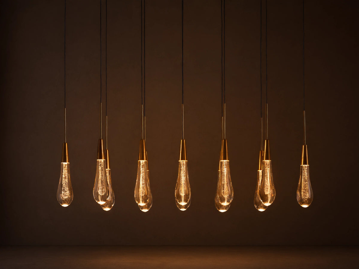 Soltaire Linear Chandelier with 8 clear, tear-drop glass pendants featuring brass tops & internal bubbles, glowing warmly.