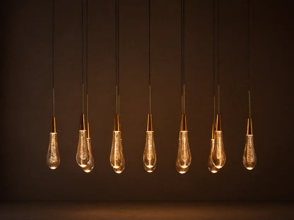 Soltaire Linear Chandelier with 8 clear, tear-drop glass pendants featuring brass tops & internal bubbles, glowing warmly.