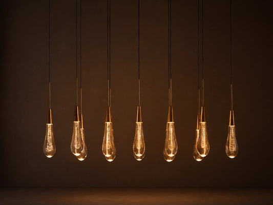 Soltaire Linear Chandelier with 8 clear, tear-drop glass pendants featuring brass tops & internal bubbles, glowing warmly.