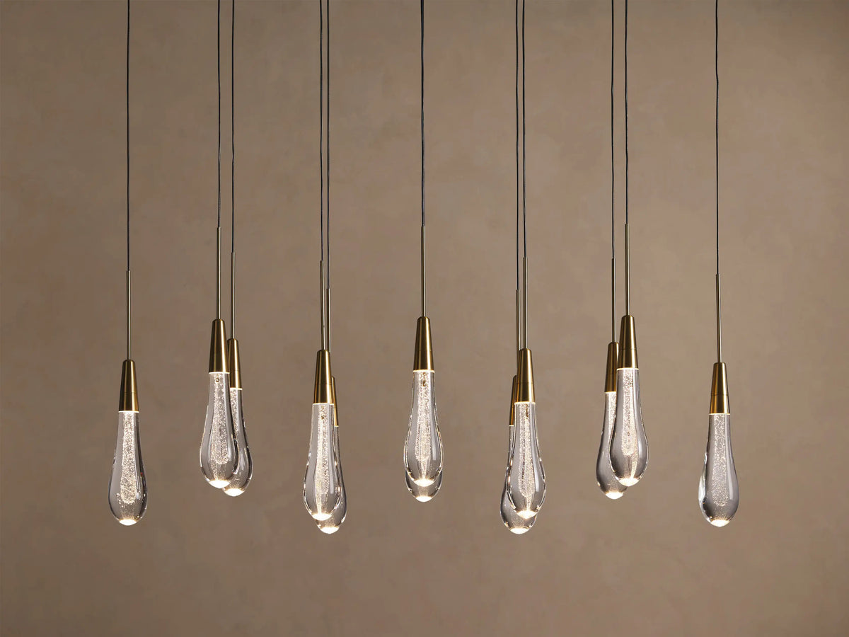 Soltaire Linear Chandelier with 10 illuminated teardrop clear glass pendants, brass-capped, hanging linearly against a warm wall.