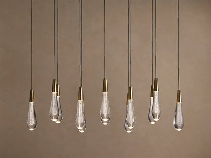 Soltaire Linear Chandelier with 10 illuminated teardrop clear glass pendants, brass-capped, hanging linearly against a warm wall.