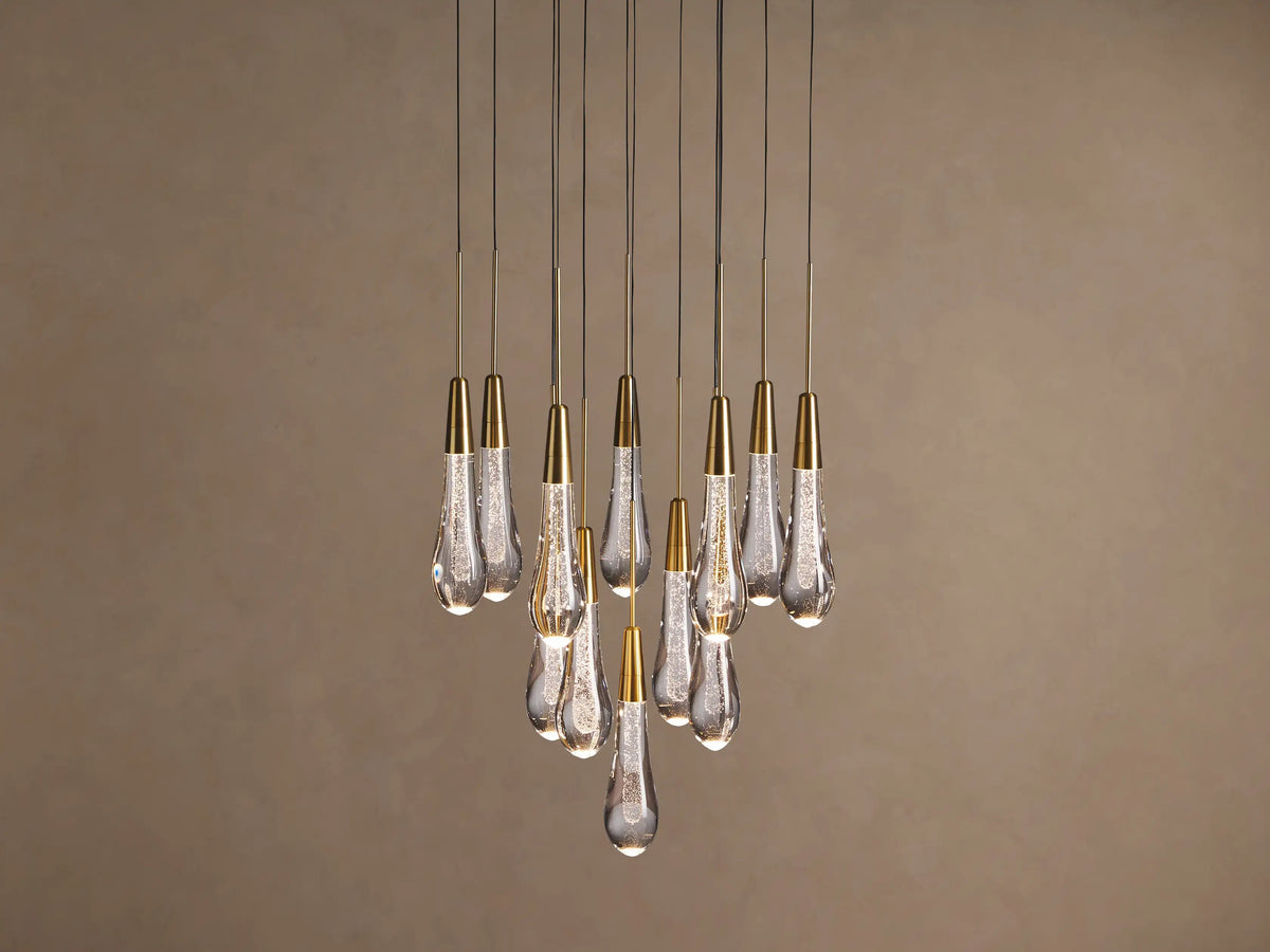 Soltaire chandelier with many illuminated teardrop glass pendants, brass caps, and internal bubbles, clustered against a neutral wall.