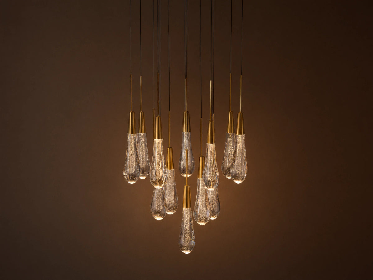 Soltaire chandelier with many illuminated teardrop glass pendants, brass caps, and internal bubbles, clustered against a neutral wall.