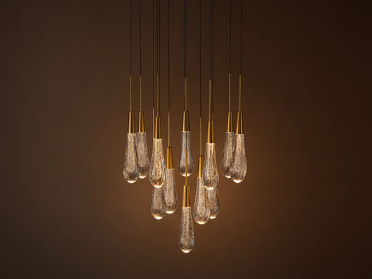 Soltaire chandelier with many illuminated teardrop glass pendants, brass caps, and internal bubbles, clustered against a neutral wall.