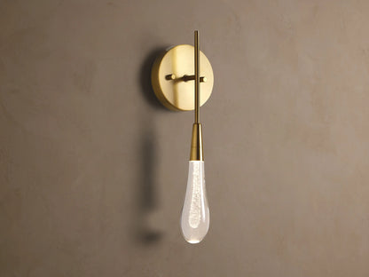 Soltaire Sconce: Round brass backplate, vertical arm, and glowing teardrop glass shade with internal bubbles mounted on a wall.