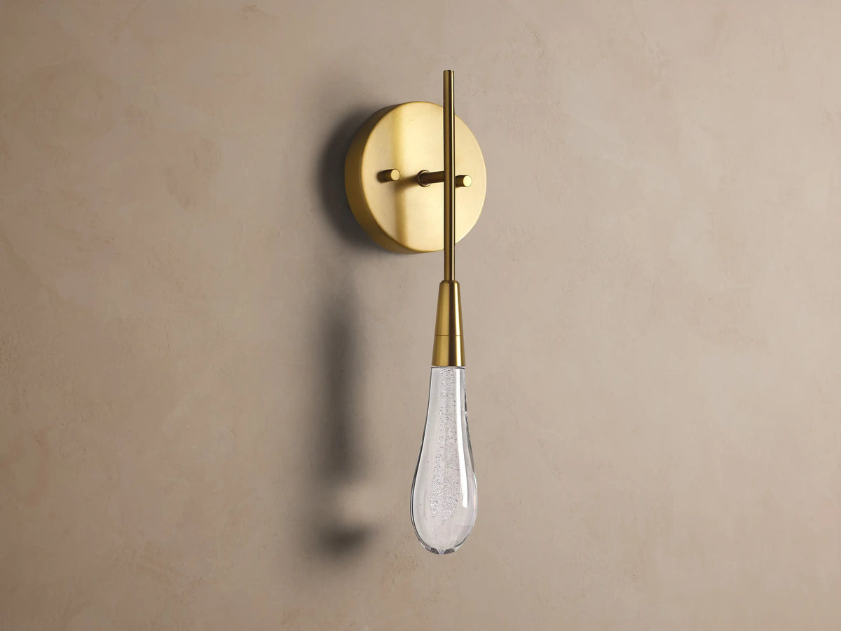 Soltaire Sconce: Round brass backplate, vertical arm, and glowing teardrop glass shade with internal bubbles mounted on a wall.
