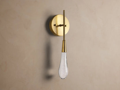 Soltaire Sconce: Round brass backplate, vertical arm, and glowing teardrop glass shade with internal bubbles mounted on a wall.