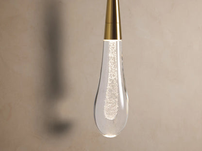 Soltaire Sconce: Round brass wall plate with a vertical rod and a clear, bubble-filled glass teardrop shade, mounted on a textured wall.
