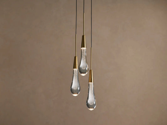 Three Soltaire Cluster Pendants with brass conical tops and clear, bubbly glass teardrop shades hang illuminated against a tan wall.