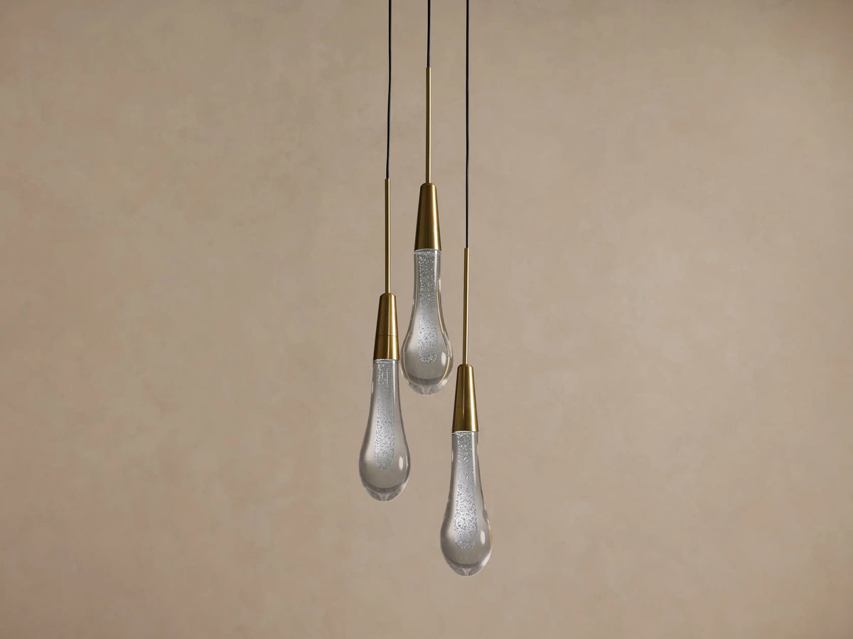Three Soltaire Cluster Pendants with brass conical tops and clear, bubbly glass teardrop shades hang illuminated against a tan wall.
