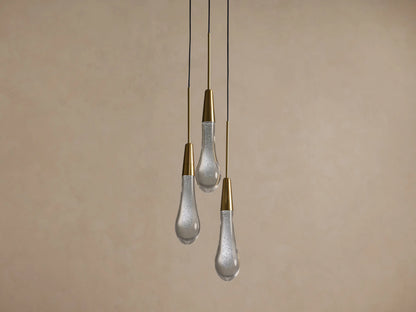 Three Soltaire Cluster Pendants with brass conical tops and clear, bubbly glass teardrop shades hang illuminated against a tan wall.