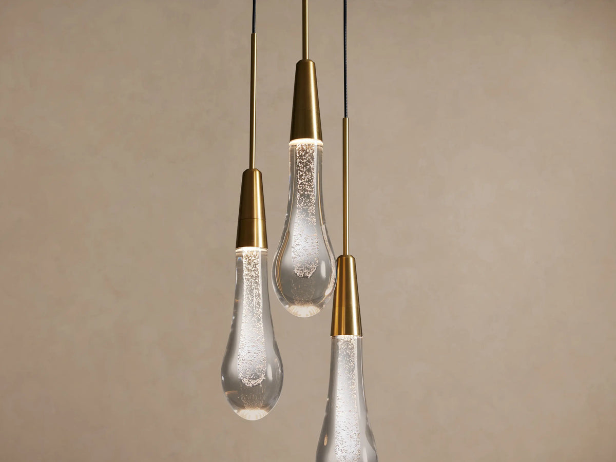 Three Soltaire Cluster Pendants with brass accents and clear tear-drop glass shades featuring internal bubbles, on a beige wall.