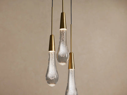 Three Soltaire Cluster Pendants with brass accents and clear tear-drop glass shades featuring internal bubbles, on a beige wall.
