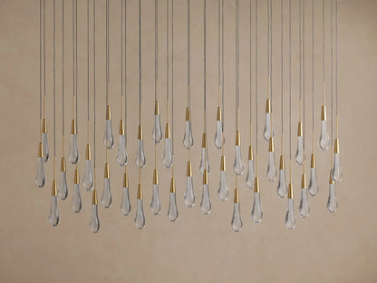 Soltaire Linear Chandelier with many clear teardrop glass pendants, brass caps, hanging at varying heights on a beige background.