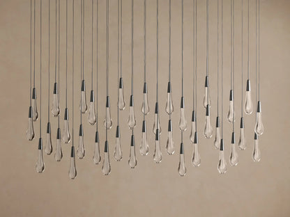 Soltaire Linear Chandelier with numerous clear tear-drop glass pendants, dark caps, suspended at varying heights against a light wall.