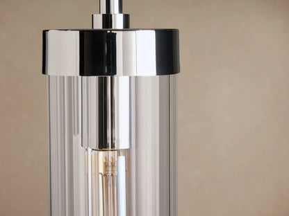 Polished nickel Andreas Pendant, featuring a clear glass cylinder shade enclosing an Edison filament bulb, suspended against a light brown wall.