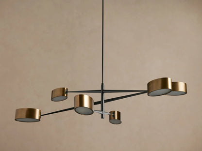 Etoile Chandelier: Modern design with black arms and six brass cylindrical shades glowing warmly, suspended against a brown wall.