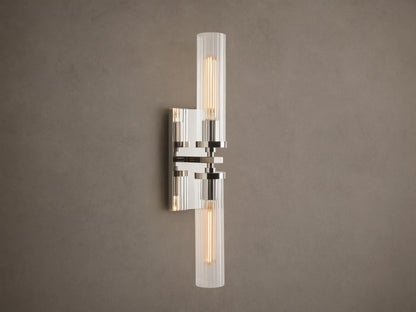 Andreas Double Sconce: vertical polished nickel hardware, two ribbed clear glass shades, warm glowing bulbs, mounted on a taupe wall.