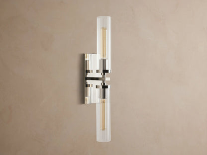 Andreas Double Sconce: vertical polished nickel hardware, two ribbed clear glass shades, warm glowing bulbs, mounted on a taupe wall.