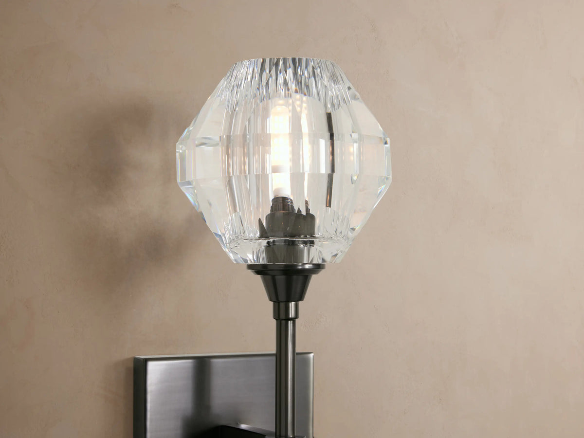 Amoroso Sconce, dark metal square backplate & long arm holding a faceted clear crystal shade, mounted on a light wall.
