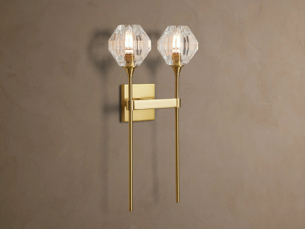 Amoroso Double Sconce, brass-plated with two illuminated faceted crystal shades, vertically mounted on a taupe wall.