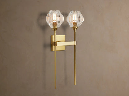 Amoroso Double Sconce, brass-plated with two illuminated faceted crystal shades, vertically mounted on a taupe wall.