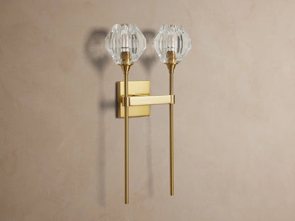 Amoroso Double Sconce, brass-plated with two illuminated faceted crystal shades, vertically mounted on a taupe wall.