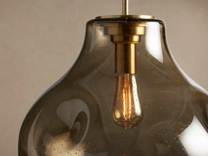 Organic 16" Pendant: Smoky gray, organic mouth-blown glass, brass hardware, & exposed Edison bulb hanging on a neutral wall.