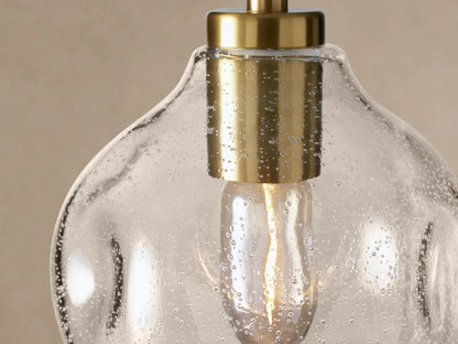 Organic 8" Pendant: Clear, seeded mouth-blown glass bulb-shaped pendant with brass fitting and black cord, against a light wall.