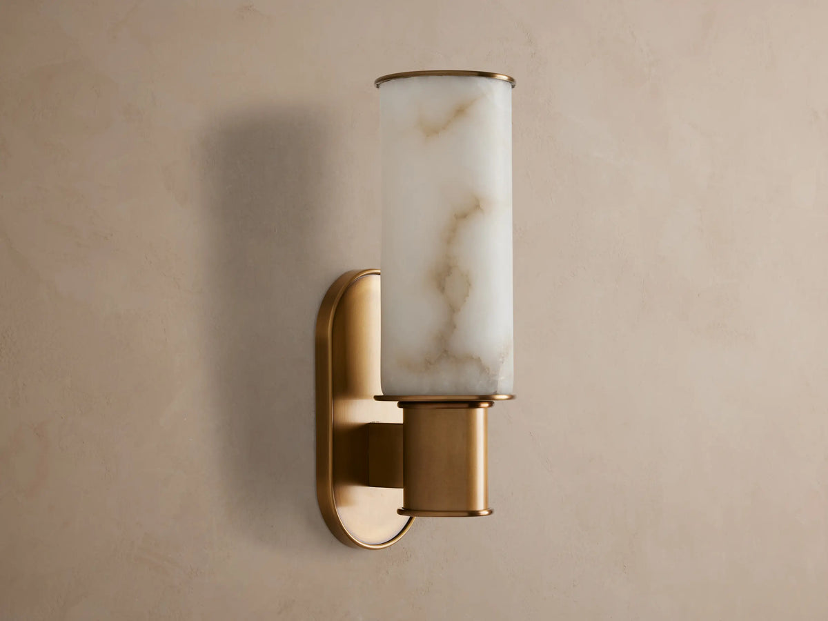 Harlin Sconce in Alabaster with a cylindrical cloche & burnished brass base, glowing warmly against a gray wall.