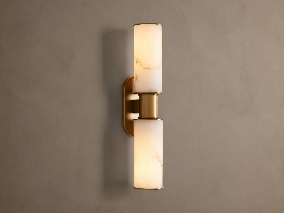 Harlin Double Sconce: Two glowing alabaster cylinder shades with burnished brass trim, mounted vertically on a neutral wall.